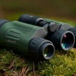 A pair of green night vision binoculars resting on mossy ground in an outdoor forest setting. The binoculars have a robust design with black rubber grips and lenses that display subtle reflections of light, indicating their optical capability.