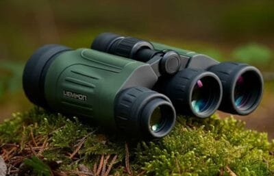 A pair of green night vision binoculars resting on mossy ground in an outdoor forest setting. The binoculars have a robust design with black rubber grips and lenses that display subtle reflections of light, indicating their optical capability.